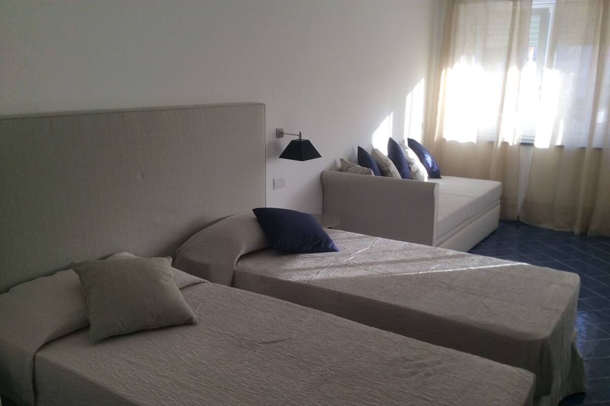 A hotel room with two single beds, a sofa bed, a bedside table, and a window with curtains.
