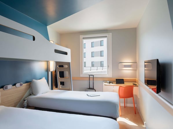 A hotel room with two beds, a desk, a chair, a television, and a window with a view of buildings. - ibis budget Saint Quentin en Yvelines Velodrome (Montigny-le-Bretonneux)