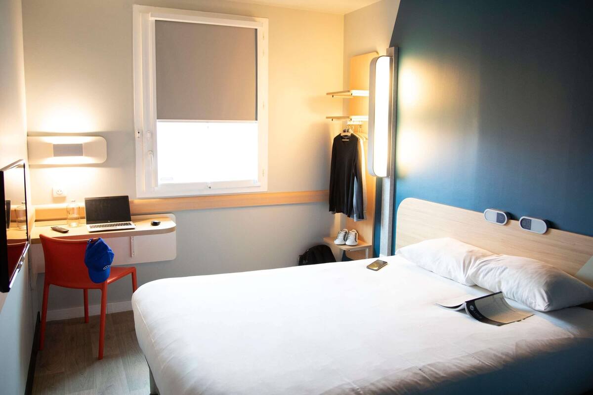 A hotel room with a bed, a desk with a laptop, a chair, a bedside table with a phone, and a window.