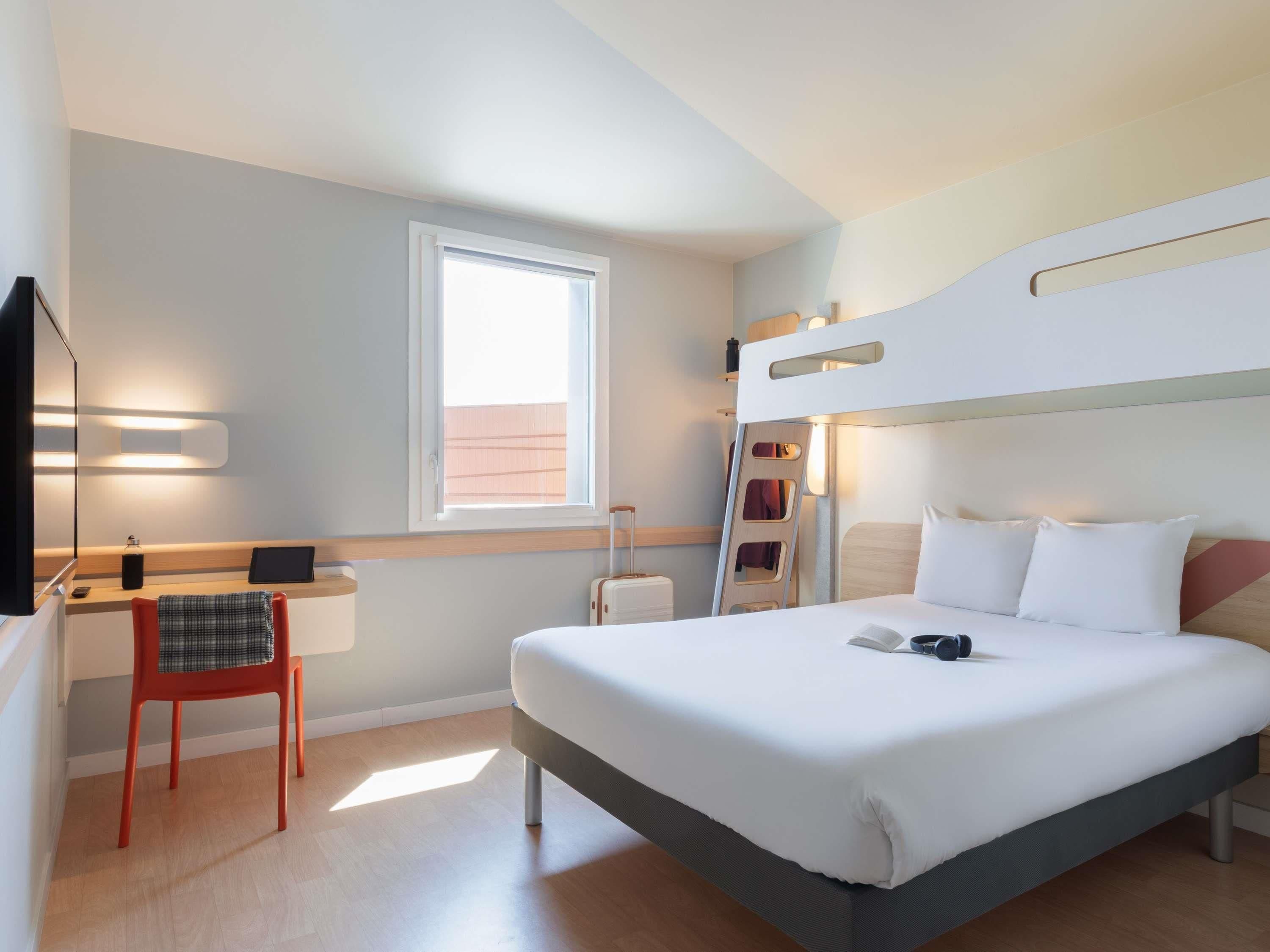A hotel room with a bunk bed, a desk with a chair, a bedside table, and a window.