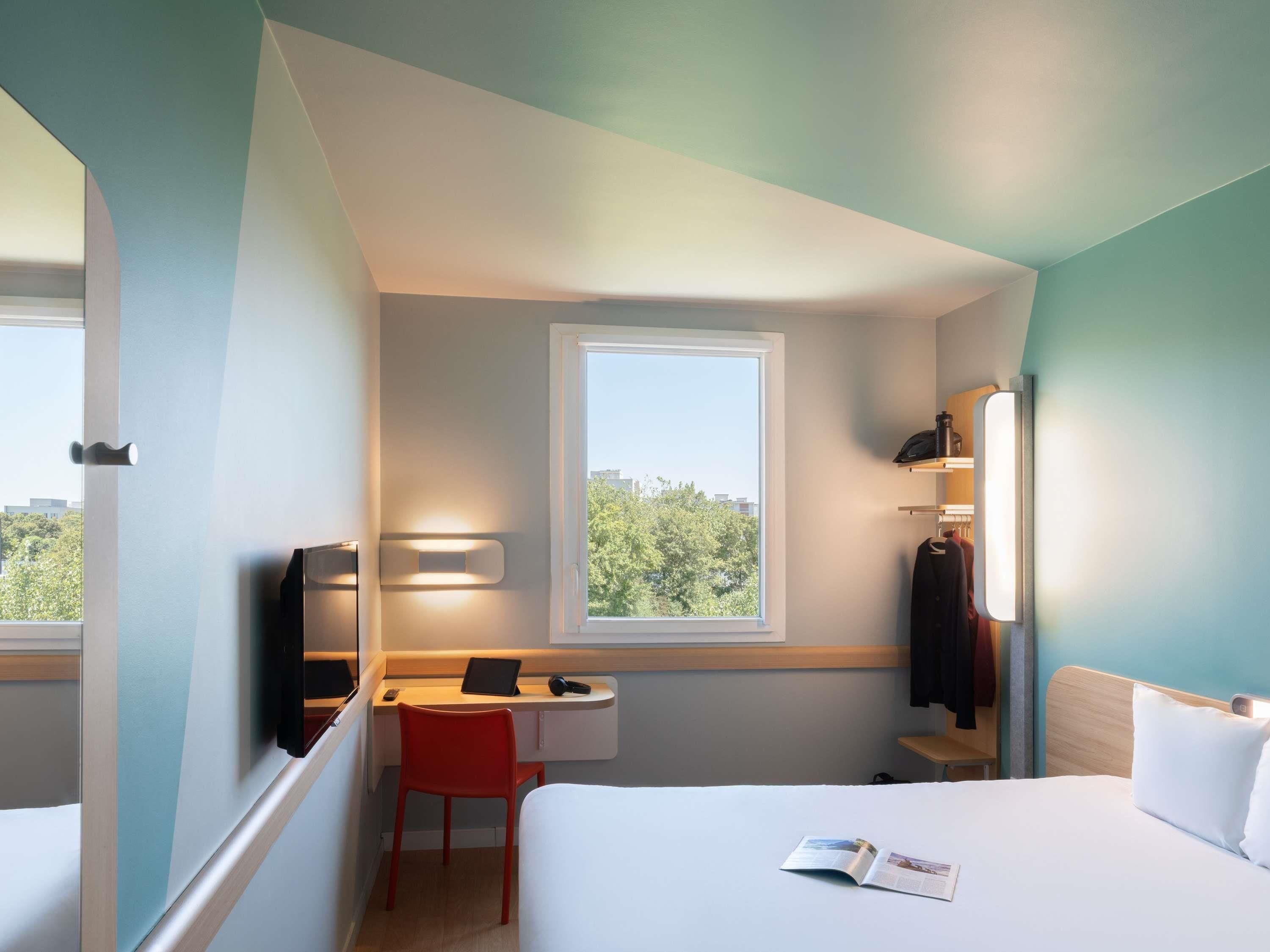 A compact hotel room with a bed, a desk with a chair, a small window, and a wardrobe.