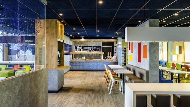 A modern cafe interior with a counter, seating area, and a view through a glass wall. - ibis budget Saint Quentin en Yvelines Velodrome (Montigny-le-Bretonneux)