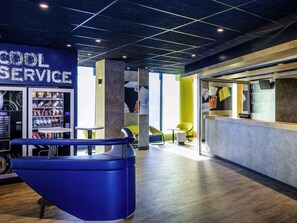 A modern interior with a vending machine labeled 'COOL SERVICE,' seating areas, and a reception desk. - ibis budget Saint Quentin en Yvelines Velodrome (Montigny-le-Bretonneux)