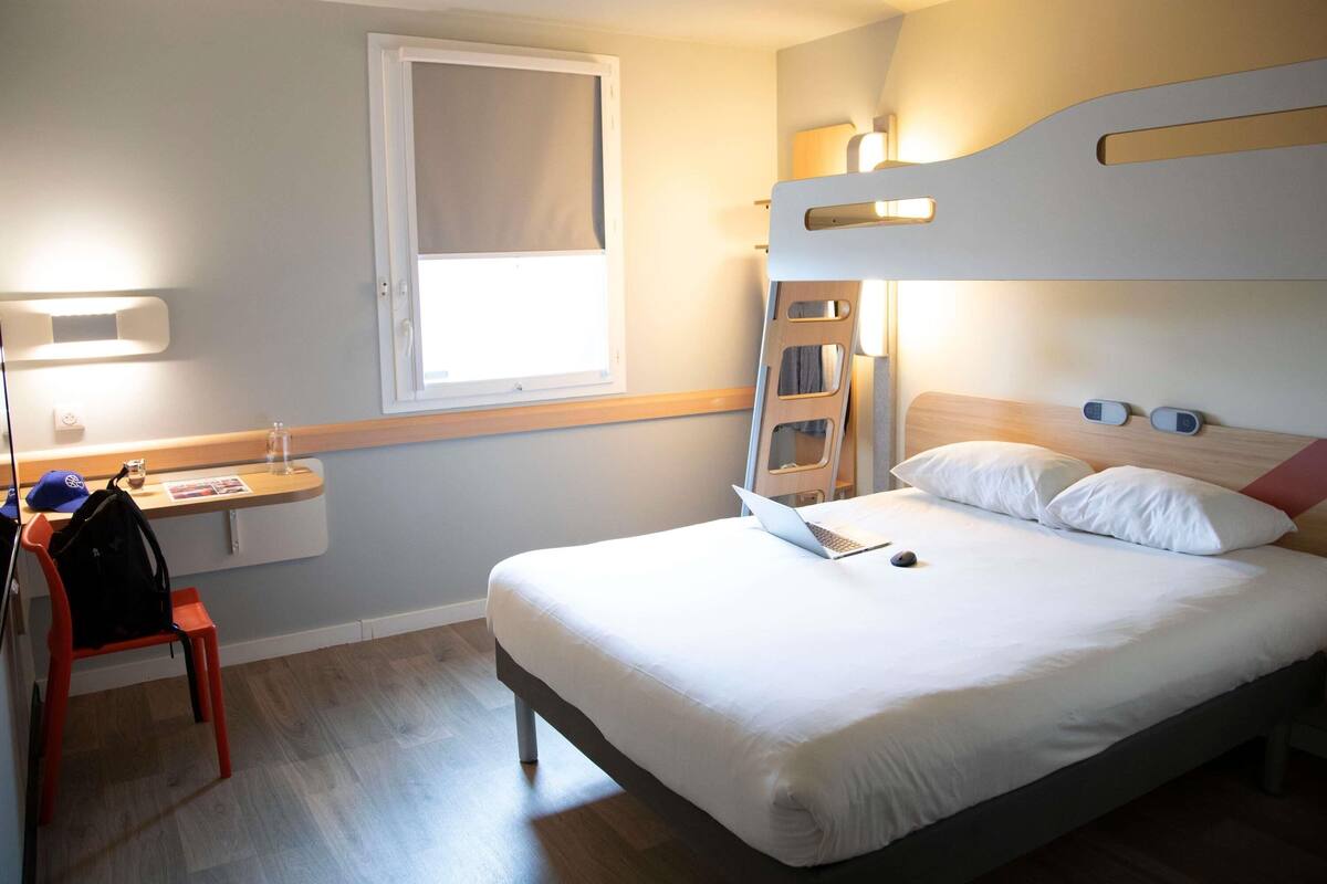 A hotel room with a bunk bed, a desk with a laptop, a chair, and a window.