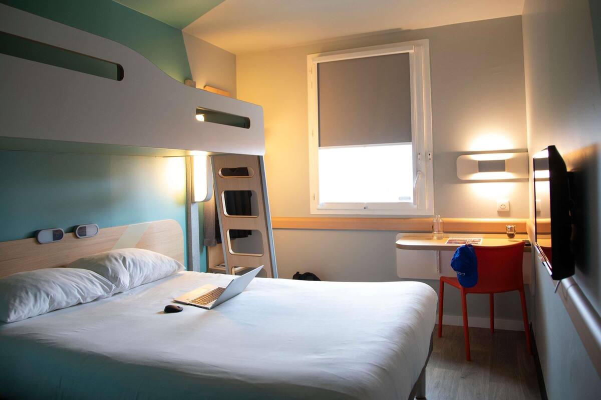 A compact hotel room with a bunk bed, a desk with a chair, a small table with a glass, and a TV.