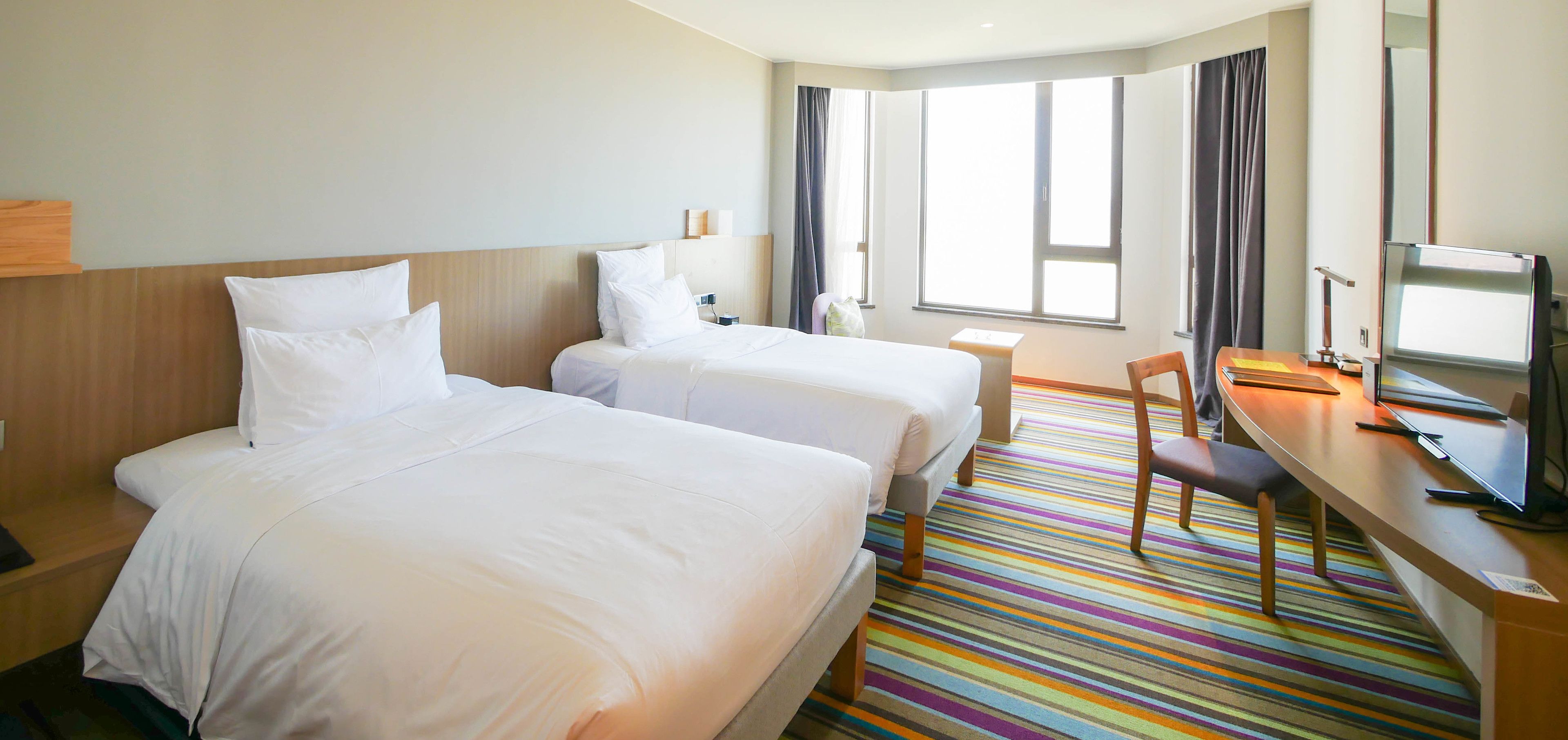 Hotellihuone, jossa on kaksi sänkyä, työpöytä, jossa on tietokone, tuoli ja suuri ikkuna.