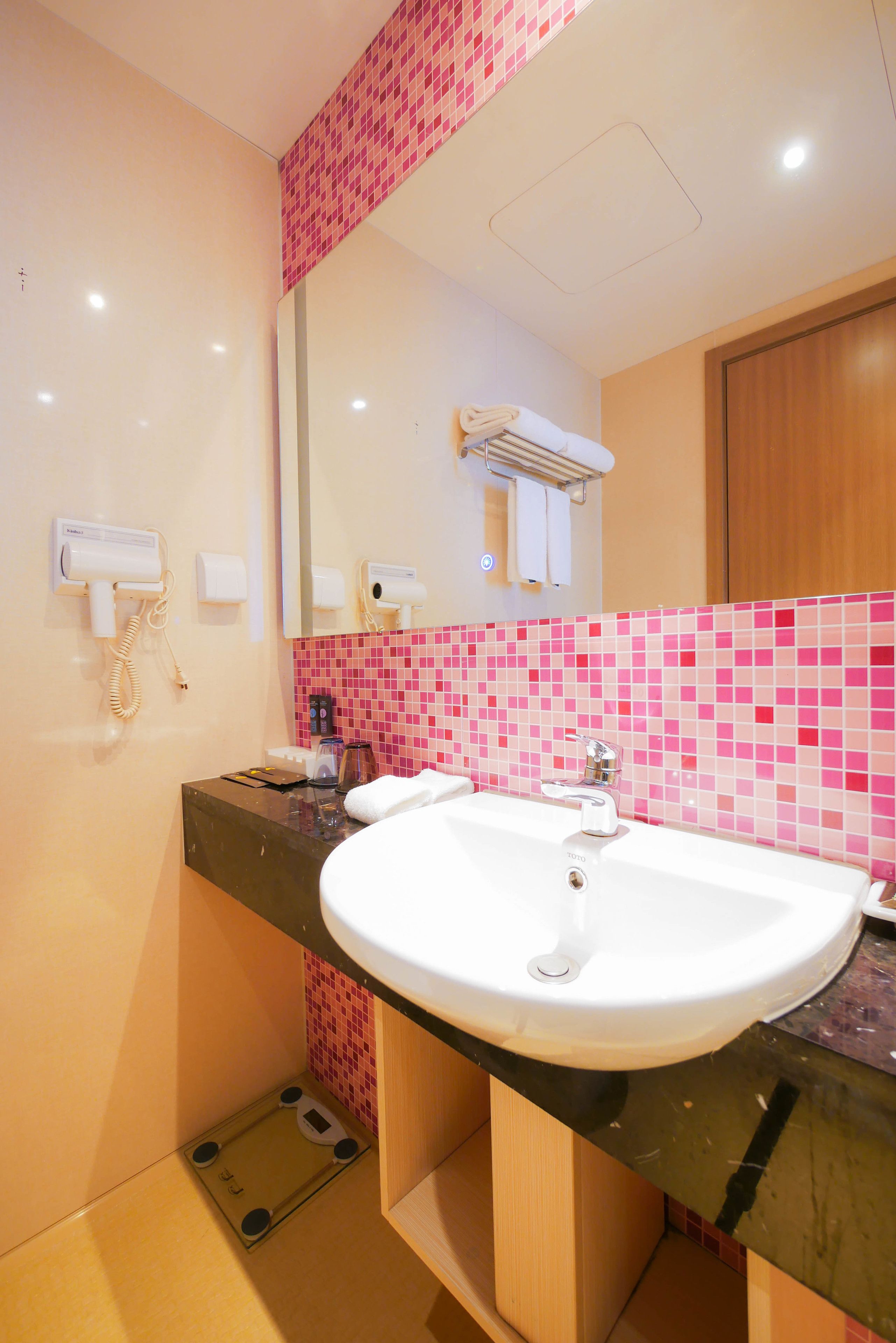 Ein Badezimmer mit einem weißen Waschbecken, rosa Mosaikfliesen, einem Spiegel, einem Föhn und einer Waage.