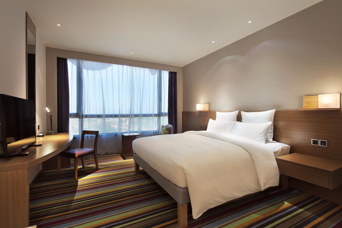 Una habitación de hotel moderna con una cama grande, un escritorio con silla, un televisor y un ventanal con cortinas.
