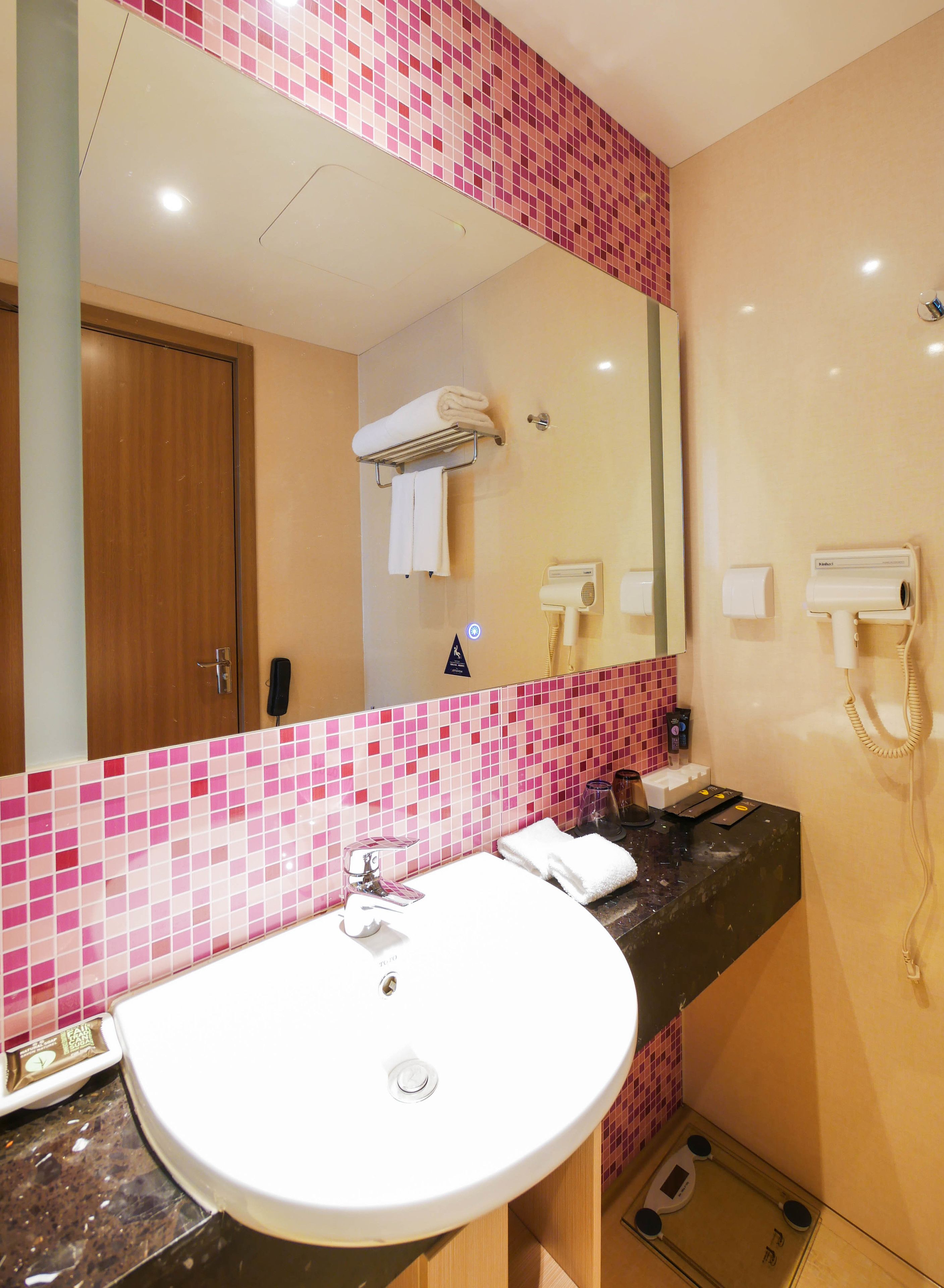 Un baño con un lavabo blanco, un espejo, baldosas de mosaico rosa, un perchero, un secador de pelo y un dispensador de papel higiénico.