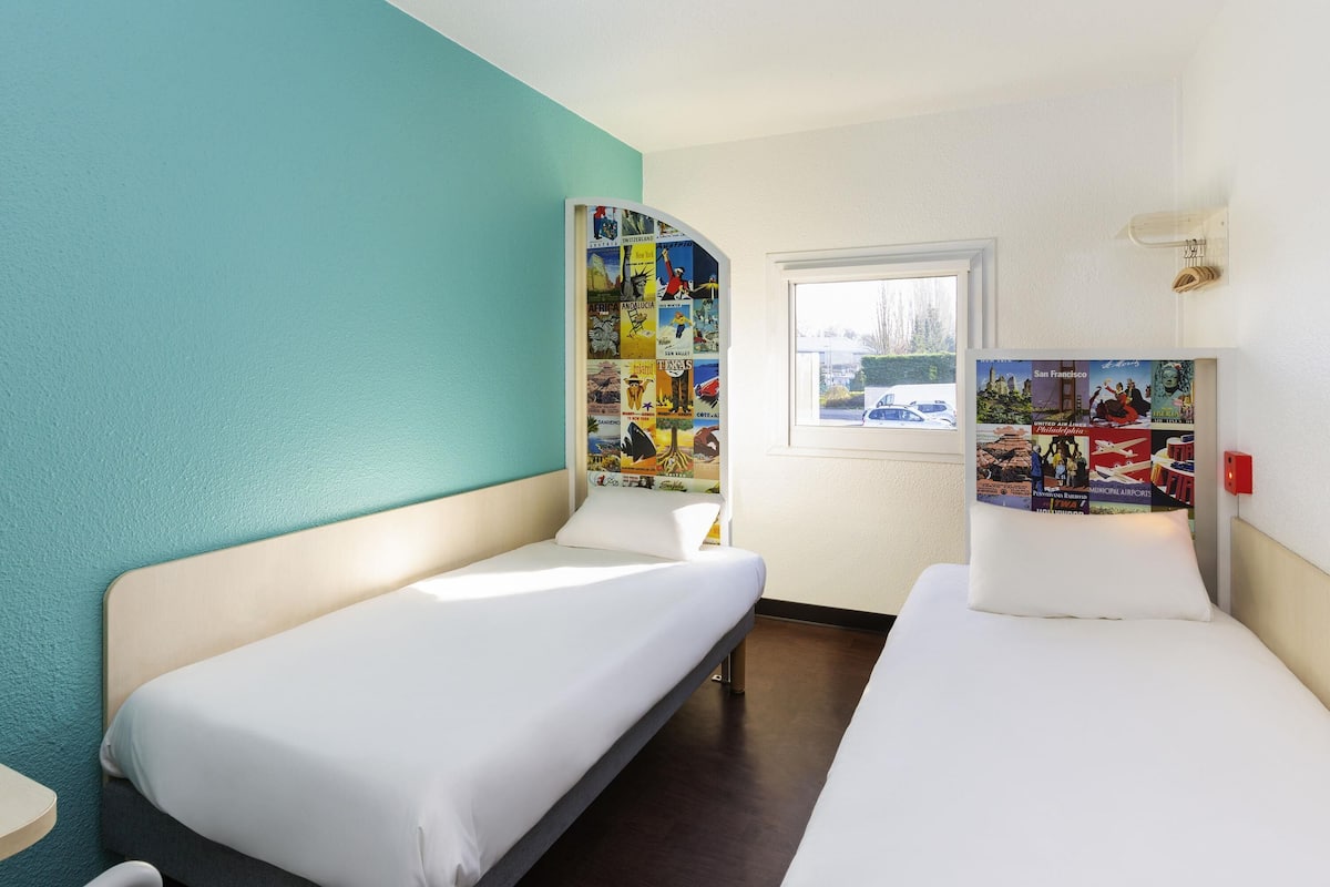 Una habitación de hotel con dos camas individuales, una ventana con vistas y una pared decorada con carteles de viajes.