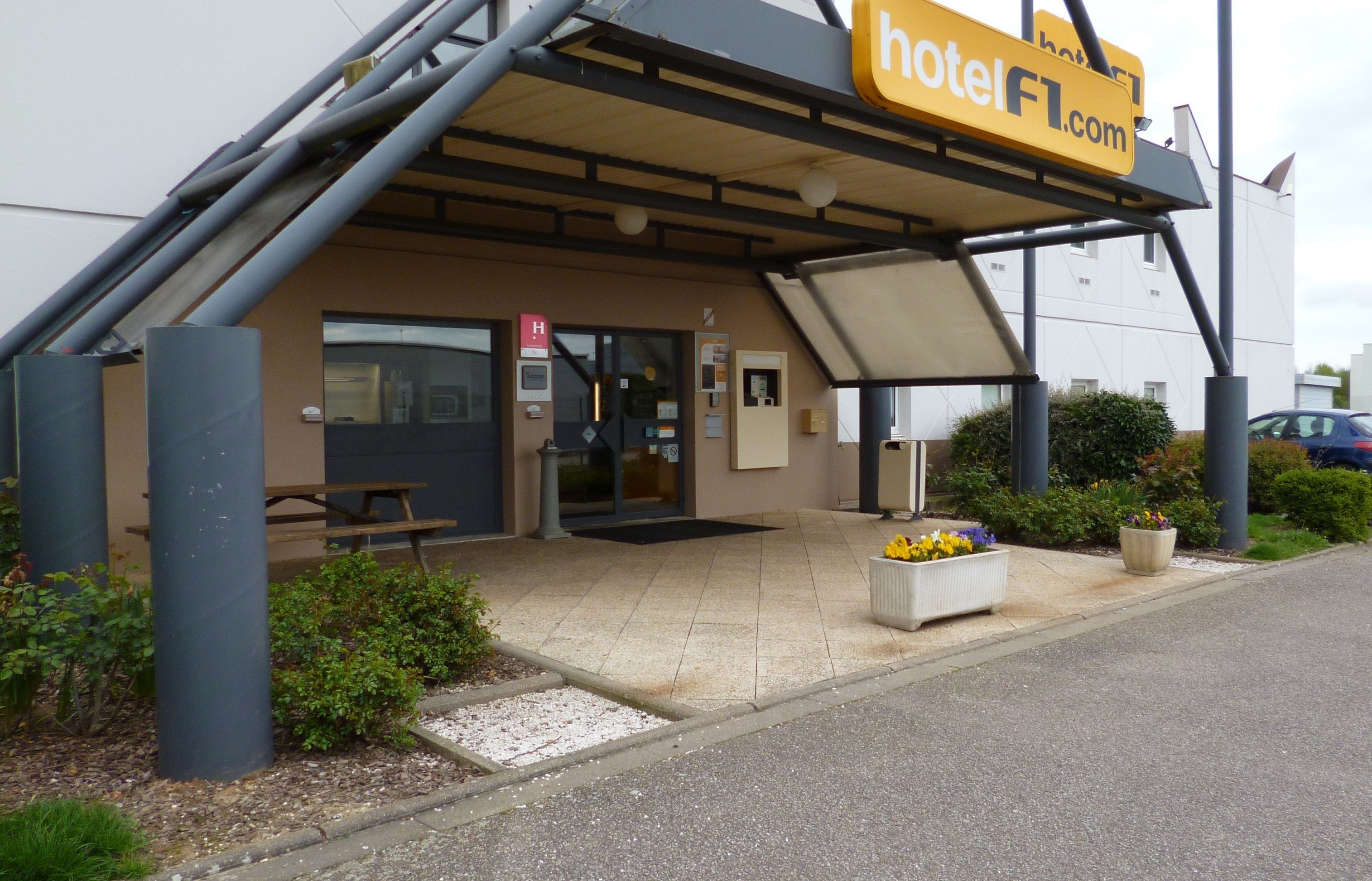 Entrada de hotel com área de estar externa coberta, porta de acesso visível e uma placa.