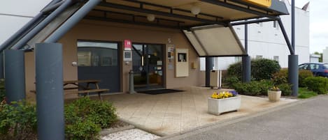 A hotel entrance with a covered outdoor seating area, a visible entrance door, and a signboard.