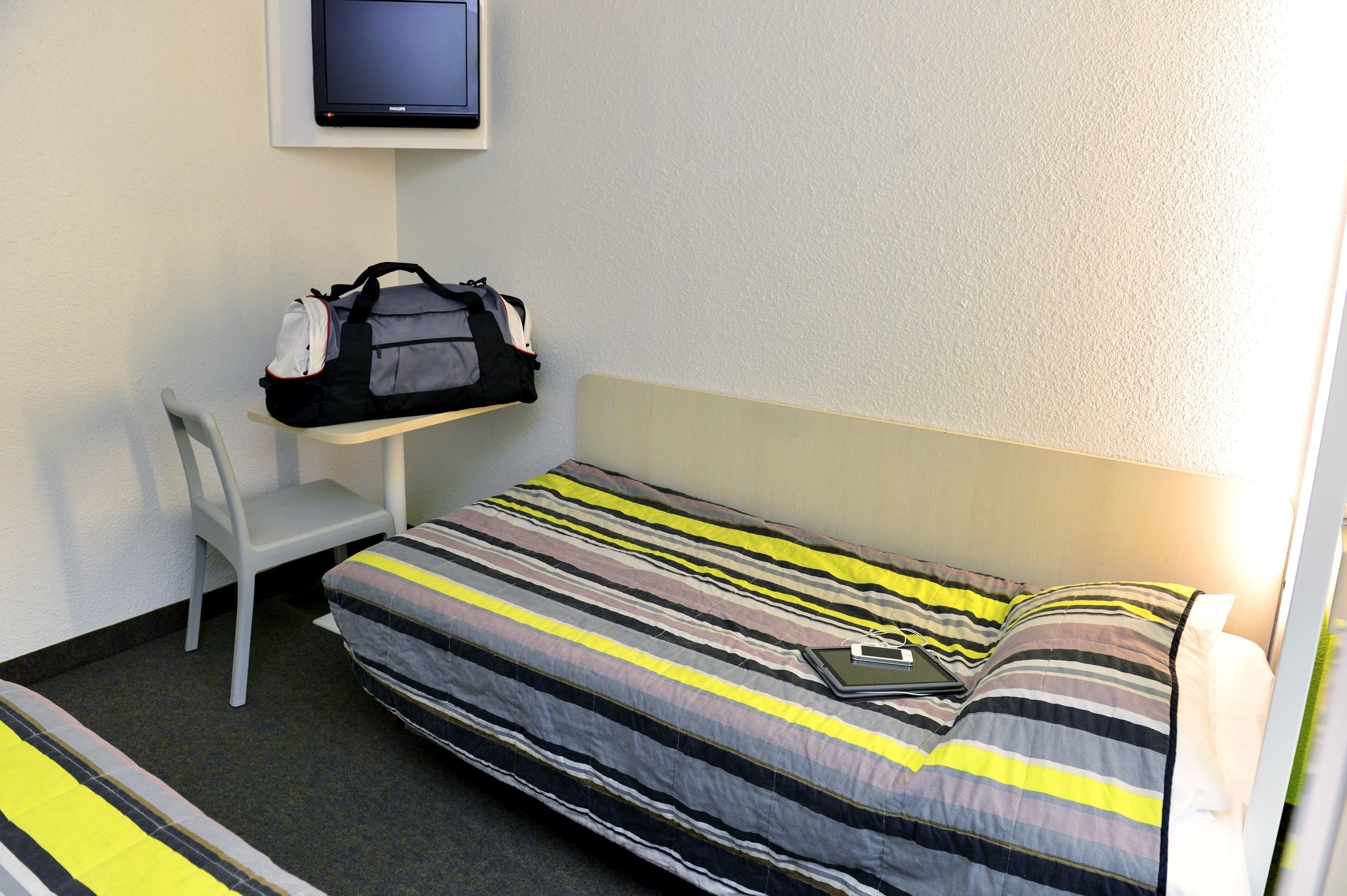 Ein Einzelbett mit gestreifter Bettdecke, ein Sessel, ein Schreibtisch mit einer Tasche und ein Fernseher an der Wand.