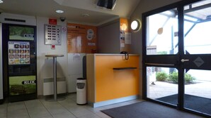 Zona de recepción de hotel con una máquina expendedora K-LIX, un cubo de basura y un mostrador de recepción.