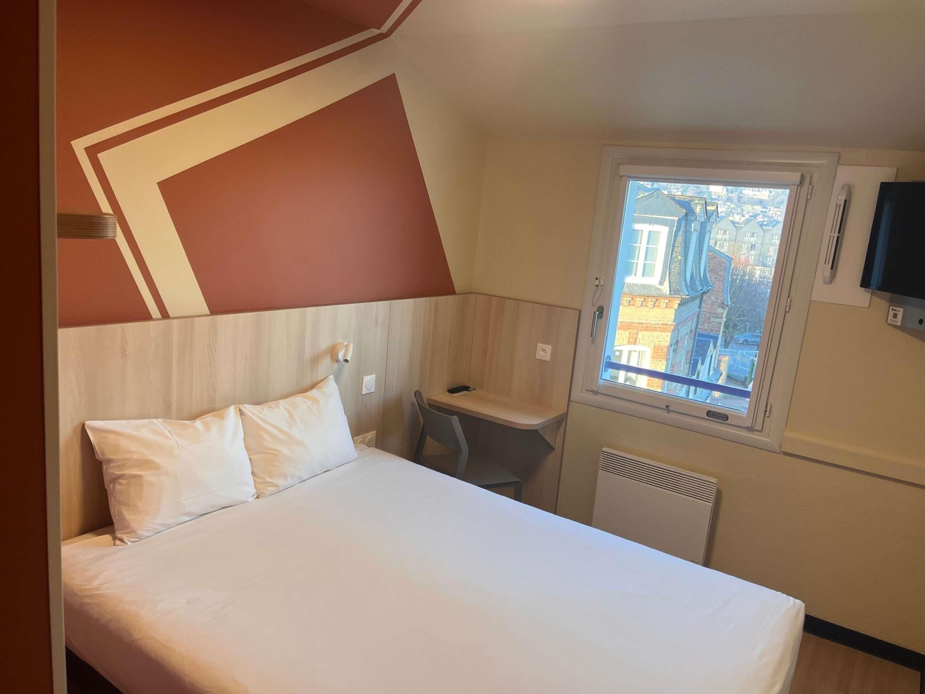 A hotel room with a bed, a small desk, a window with a view of buildings, and a TV mounted on the wall.