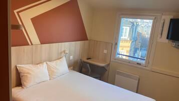 A hotel room with a bed, a small desk, a window with a view of buildings, and a TV mounted on the wall.