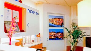 Zona de recepción con un cartel que dice 'RECEPTION', una caja registradora, folletos informativos, un florero con flores y dos cuadros enmarcados con vistas de la ciudad.