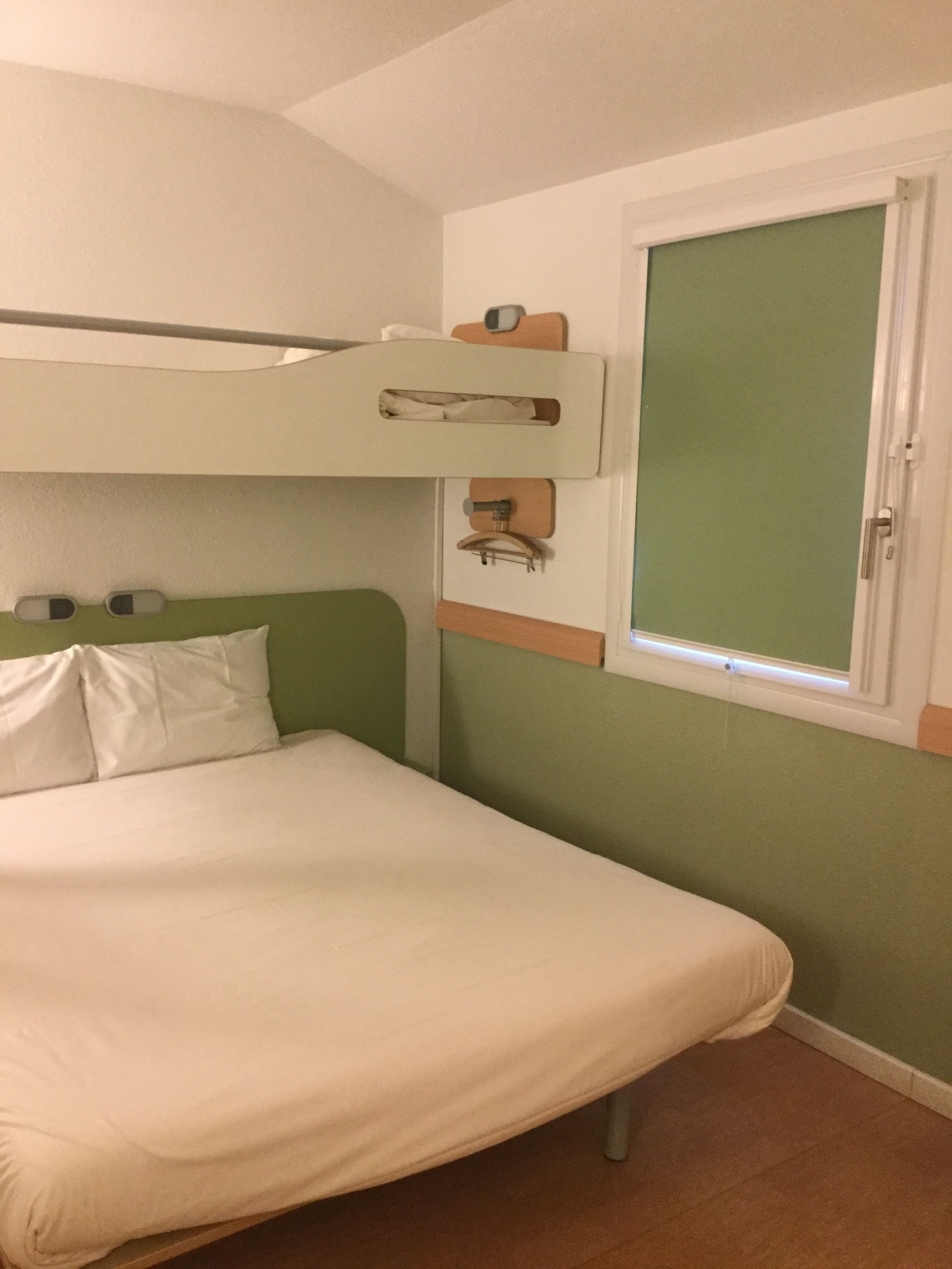 Una cama litera con cabecera verde, una cama individual en la parte inferior, una ventana con persiana verde y una puerta con marco blanco.