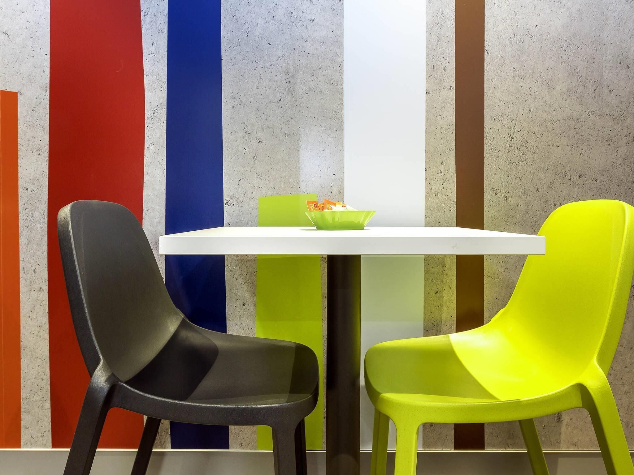 Una mesa blanca con dos sillas, una negra y otra verde, frente a una pared con rayas verticales rojas, azules y verdes.