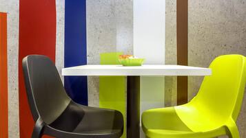 Una mesa blanca con dos sillas, una negra y otra verde, frente a una pared con rayas verticales rojas, azules y verdes.