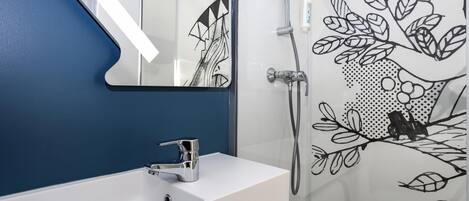 Un baño con un lavamanos blanco, un espejo, una ducha con puerta de cristal y una pared con un estampado de hojas.