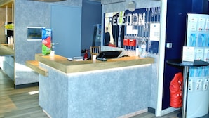 A reception desk with a counter, a fire extinguisher, a telephone, and a wall-mounted screen displaying a person. - ibis budget Flers Les Grands Champs (Flers)