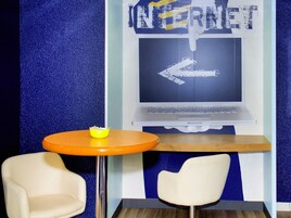 A room with a blue textured wall, a laptop on a stand, a small orange table, and two white chairs.
