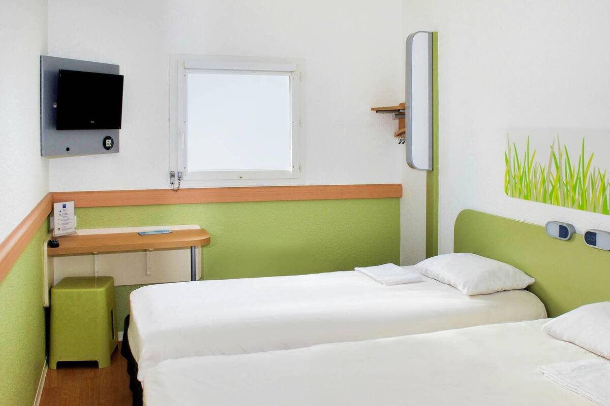 A hotel room with two beds, a small desk, a television, and a window.