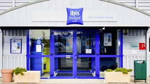 The entrance to ibis budget hotels with a blue facade and a sign indicating the location as Fleurs Les Grands Champs. - ibis budget Flers Les Grands Champs (Flers)