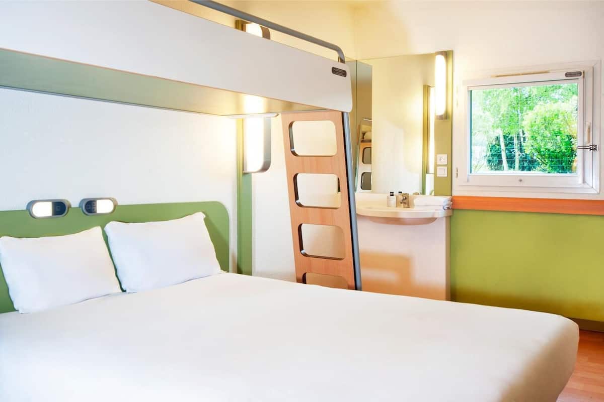 A hotel room with a bed, a bedside table, a mirror, a window, and a small bathroom area.