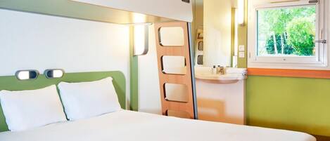 Una habitación de hotel con cama, mesita de noche, espejo, ventana y una lámpara fijada a la pared.
