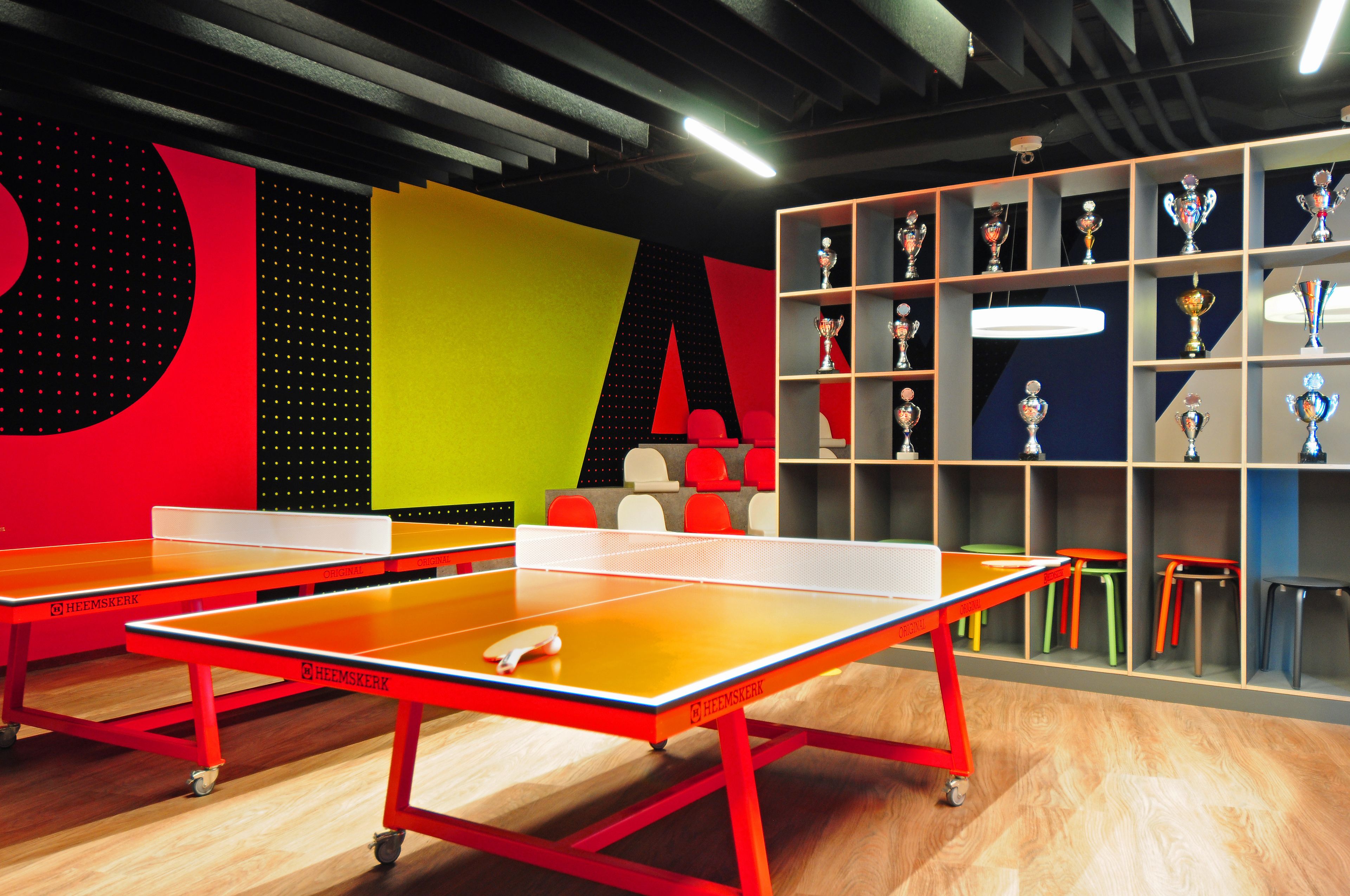 A ping pong table with a red frame and a white net, a shelf with trophies, and a colorful wall with geometric shapes.