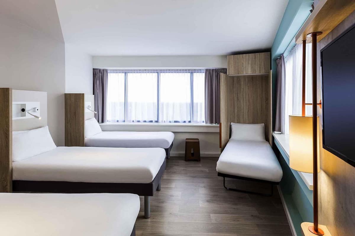 Habitación de hotel con dos camas, televisión, ventana con cortinas y un pequeño taburete.