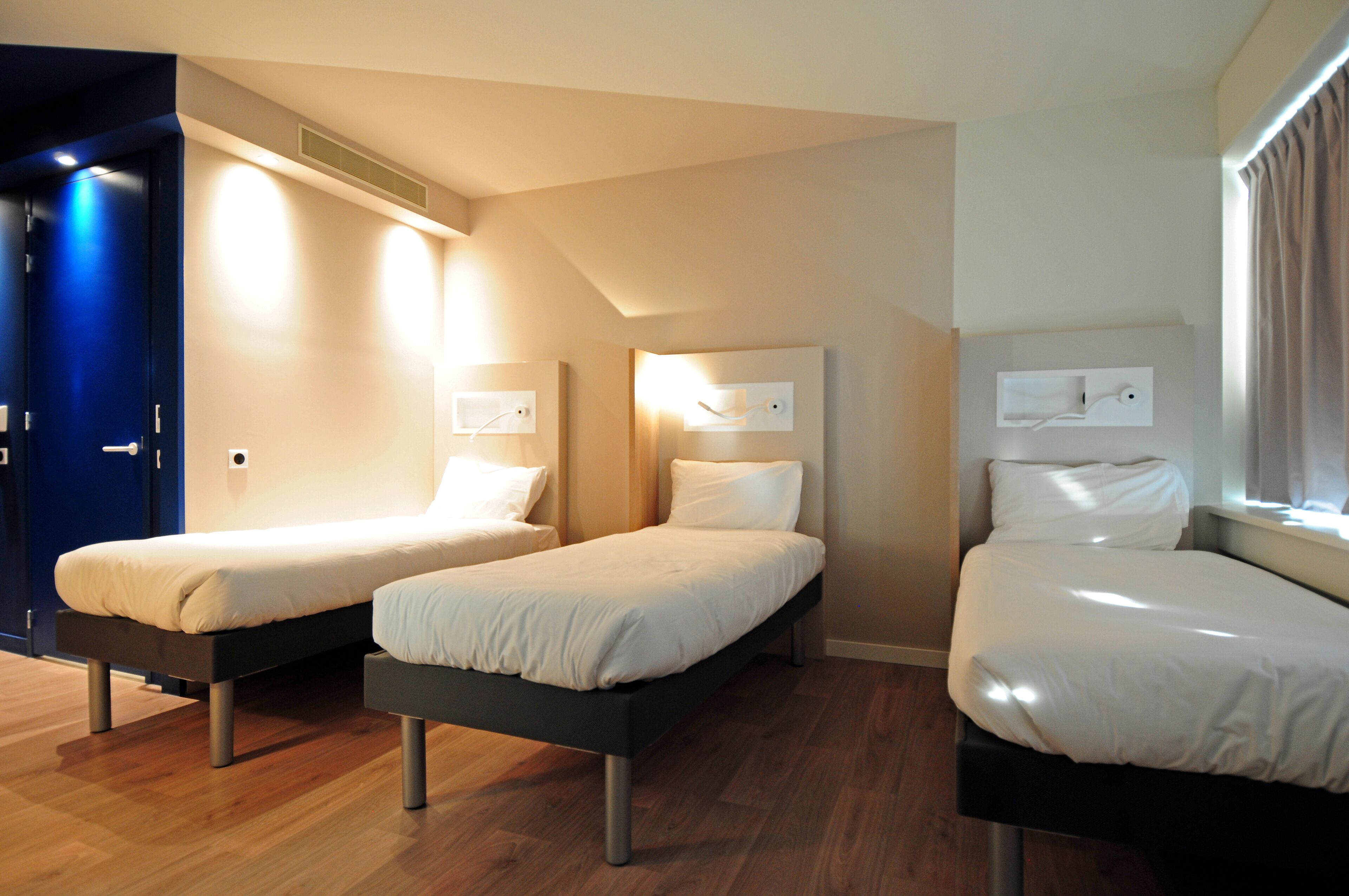 Habitación de hotel con tres camas individuales, suelo de madera y una puerta azul.