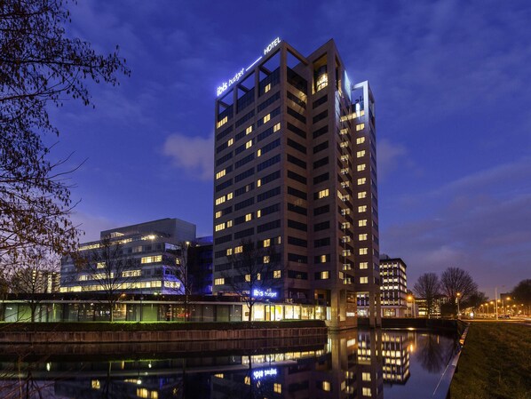 A modern hotel with a prominent 'ibis budget' sign, reflecting in a calm body of water, surrounded by urban buildings and a clear sky.