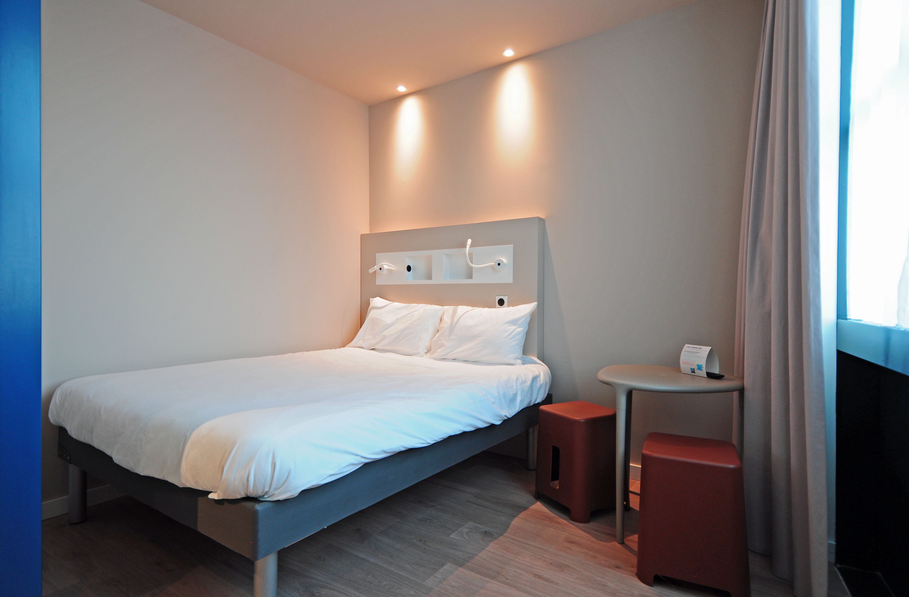 A modern hotel room with a bed, bedside table, and a small stool.