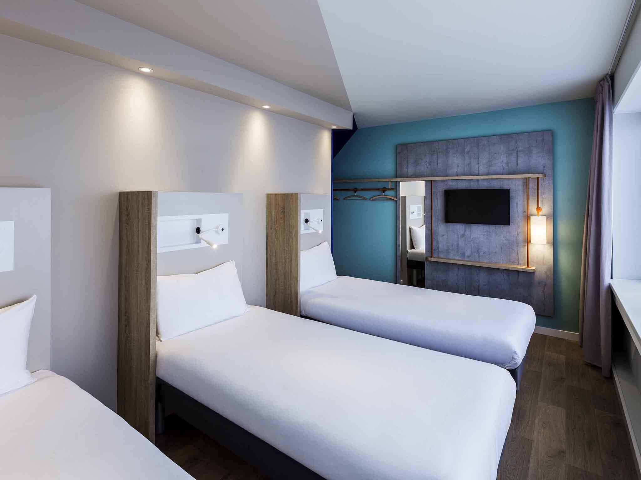 A hotel room with three single beds, a large mirror, a TV mounted on the wall, and a wooden headboard.