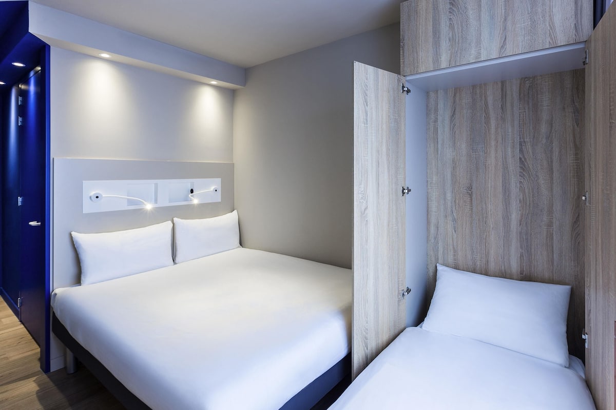 A modern hotel room with a large bed, a wardrobe, and a wall-mounted light fixture.