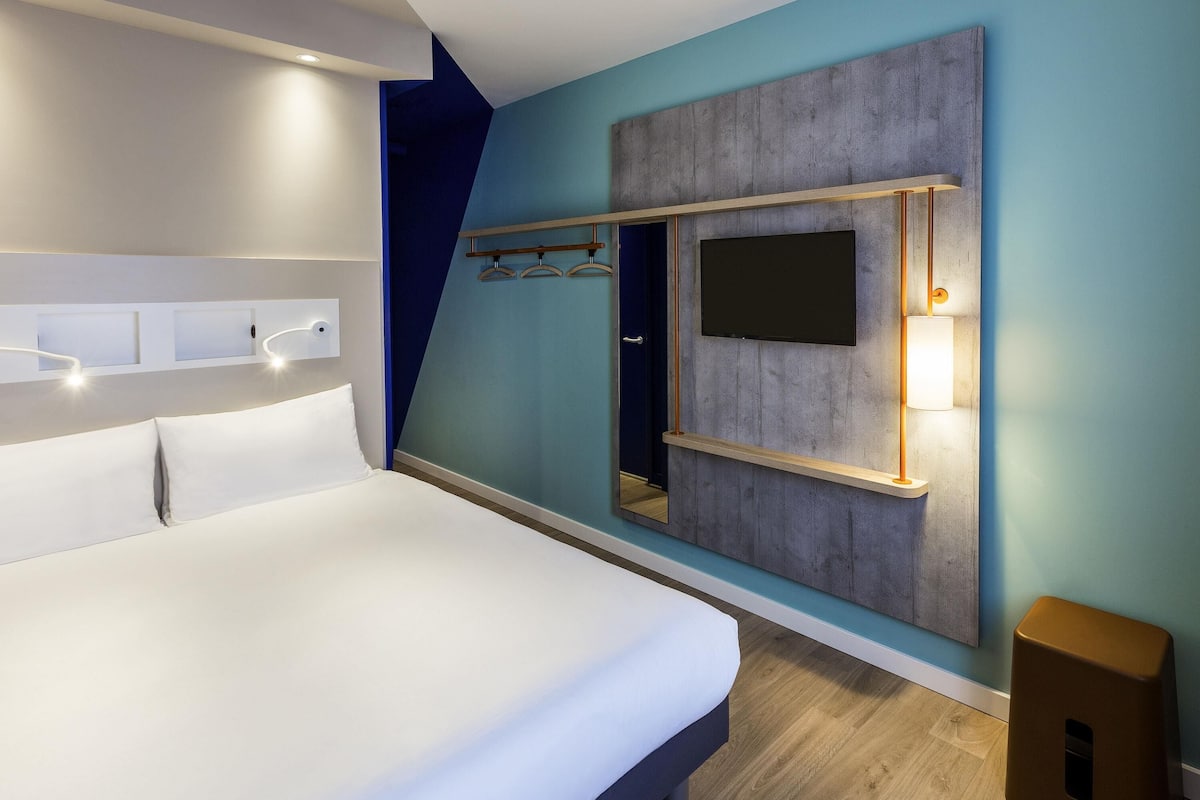 A modern hotel room with a bed, a mounted TV, and a coat rack.