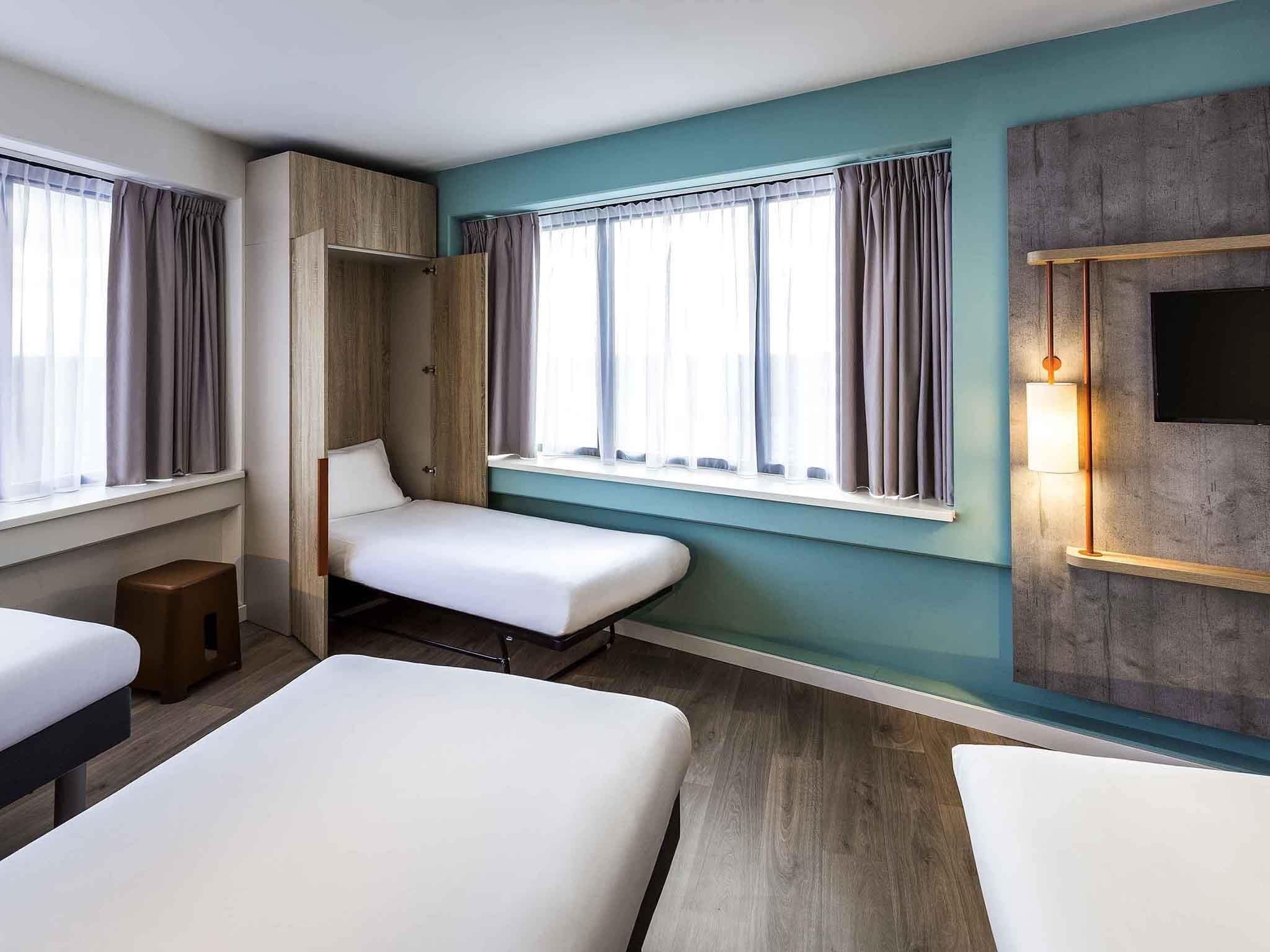 Una habitación de hotel con dos camas, un armario, un taburete pequeño y un televisor montado en la pared.