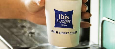 Una mano sosteniendo una taza con el texto “ibis budget for a smart start” cerca de una máquina de café.