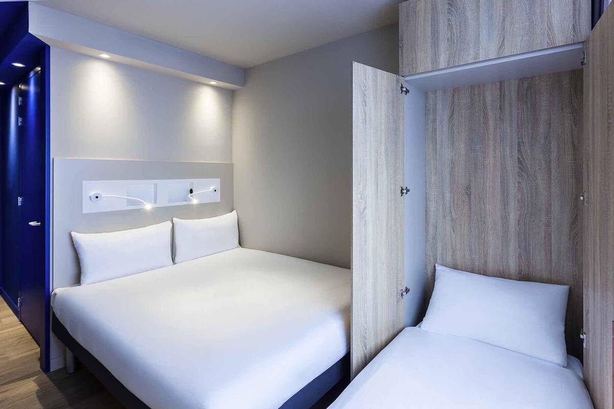Una habitación de hotel moderna con una cama grande, un armario de madera y una lámpara empotrada sobre la cama.