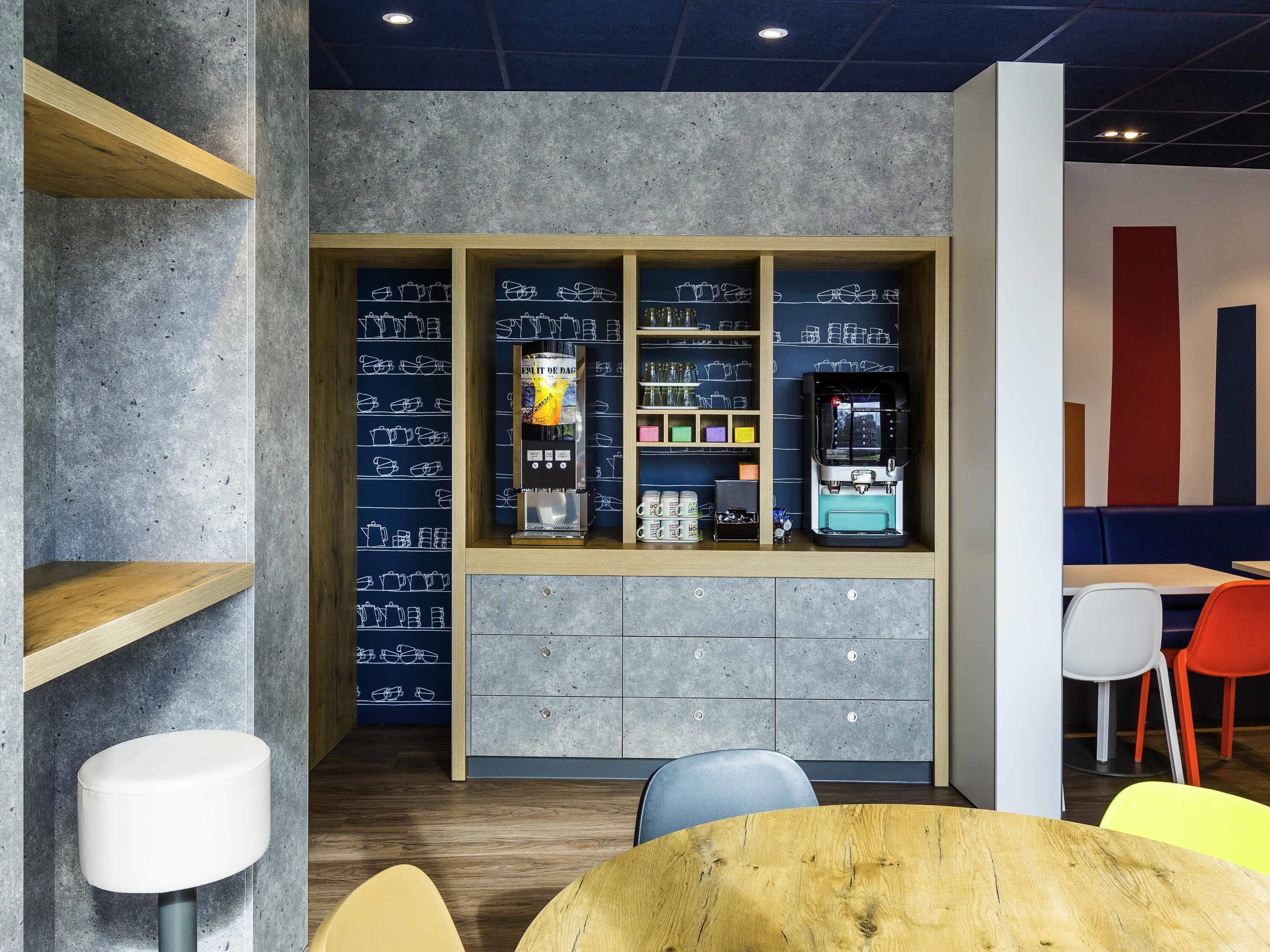 A modern interior with a coffee machine, shelves with mugs, and a seating area with colorful chairs.