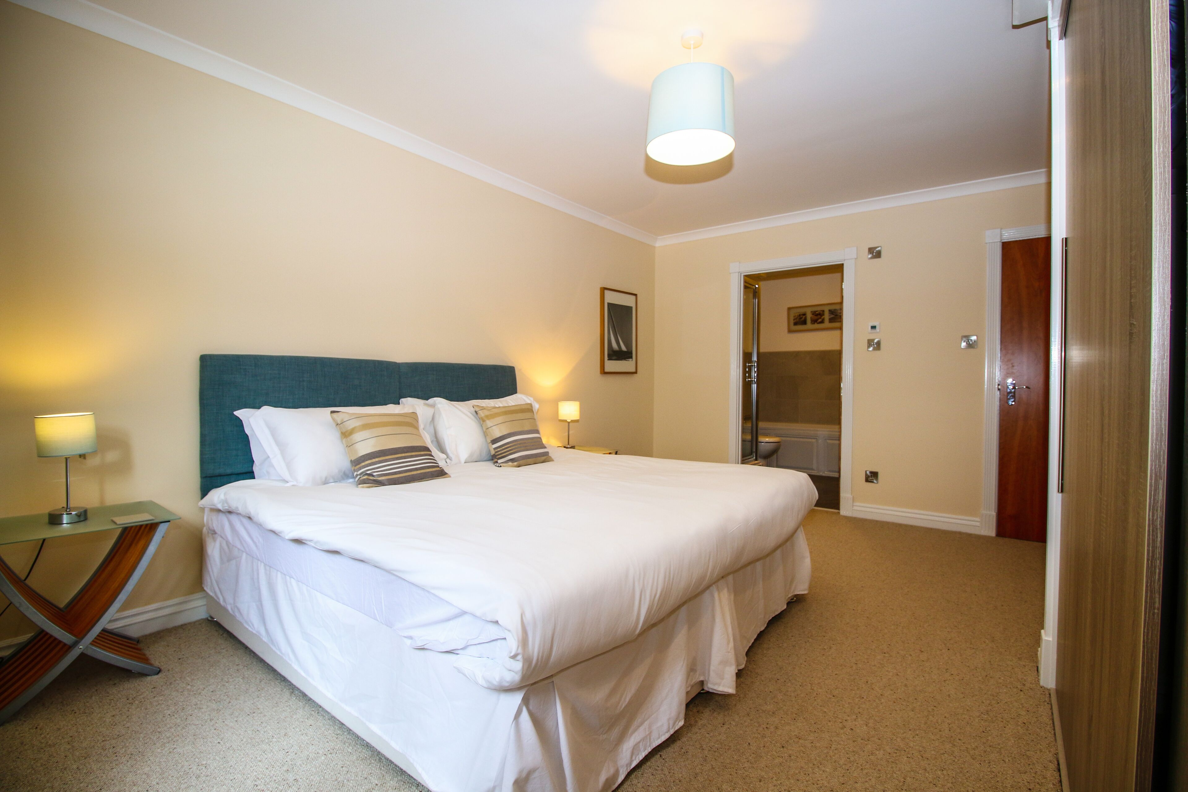 A neatly made bed with white bedding and multiple pillows, a bedside table with a lamp, and a door leading to a bathroom.