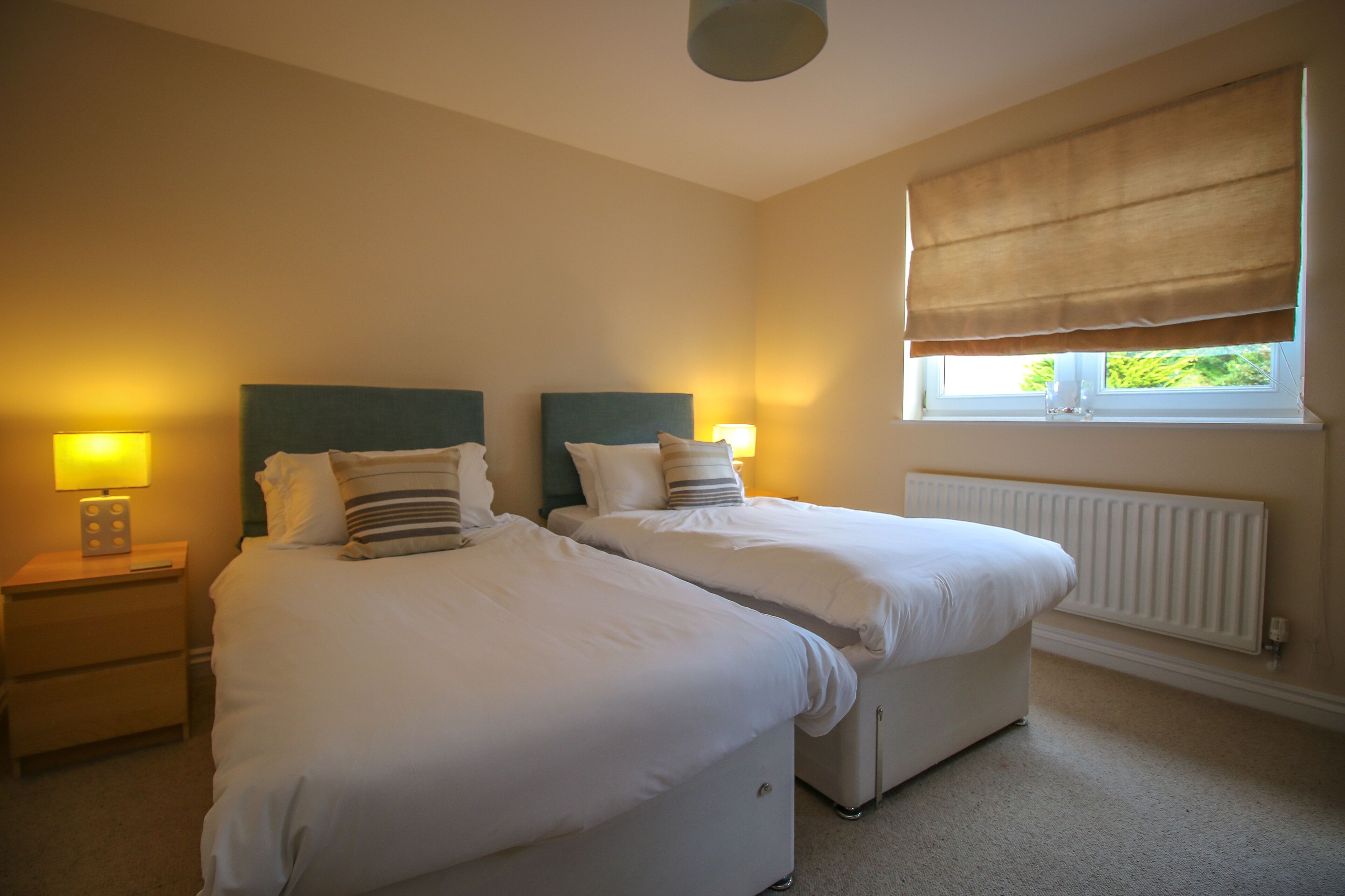 A bedroom with two single beds, bedside tables with lamps, a window with a blind, and a radiator.