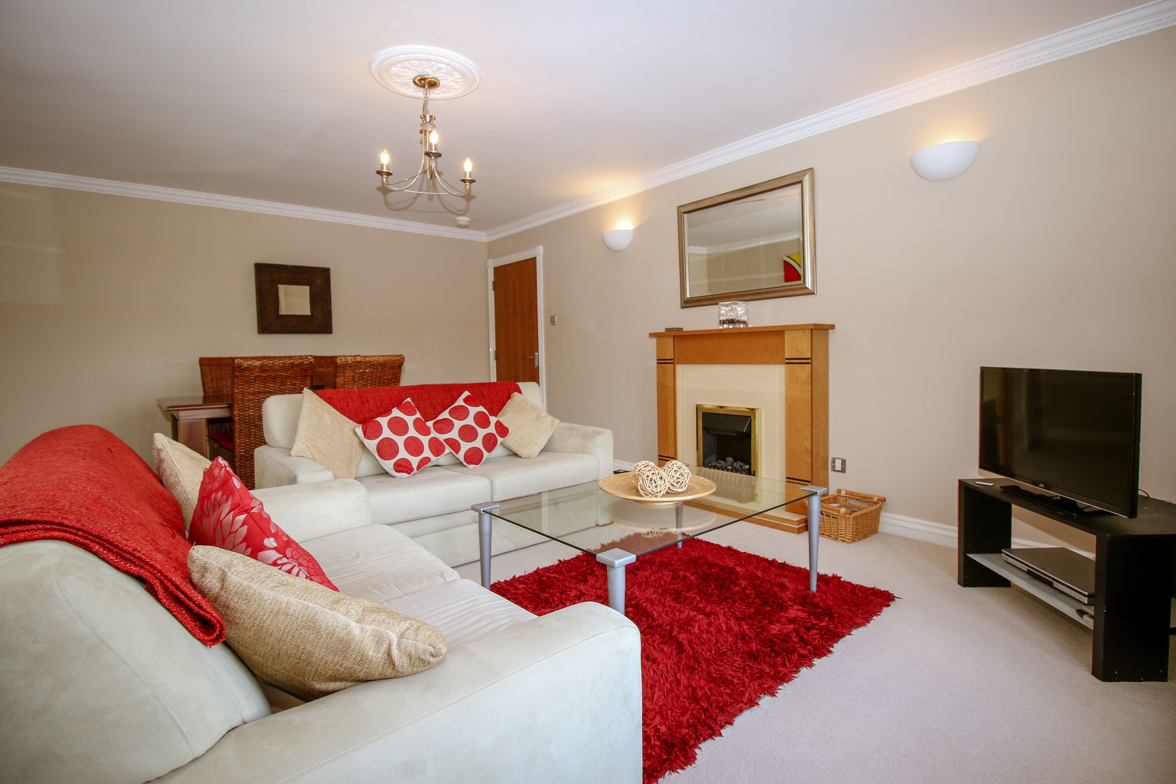 A living room with a white sofa, red and white cushions, a glass coffee table, a red rug, a fireplace, and a TV on a stand.