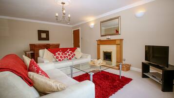 A living room with a white sofa, red and white cushions, a glass coffee table, a red rug, a fireplace, and a TV on a stand.