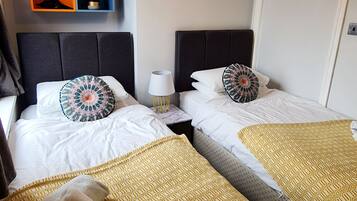 A hotel room with two beds, a small table, a lamp, and a wall-mounted shelf with potted plants.