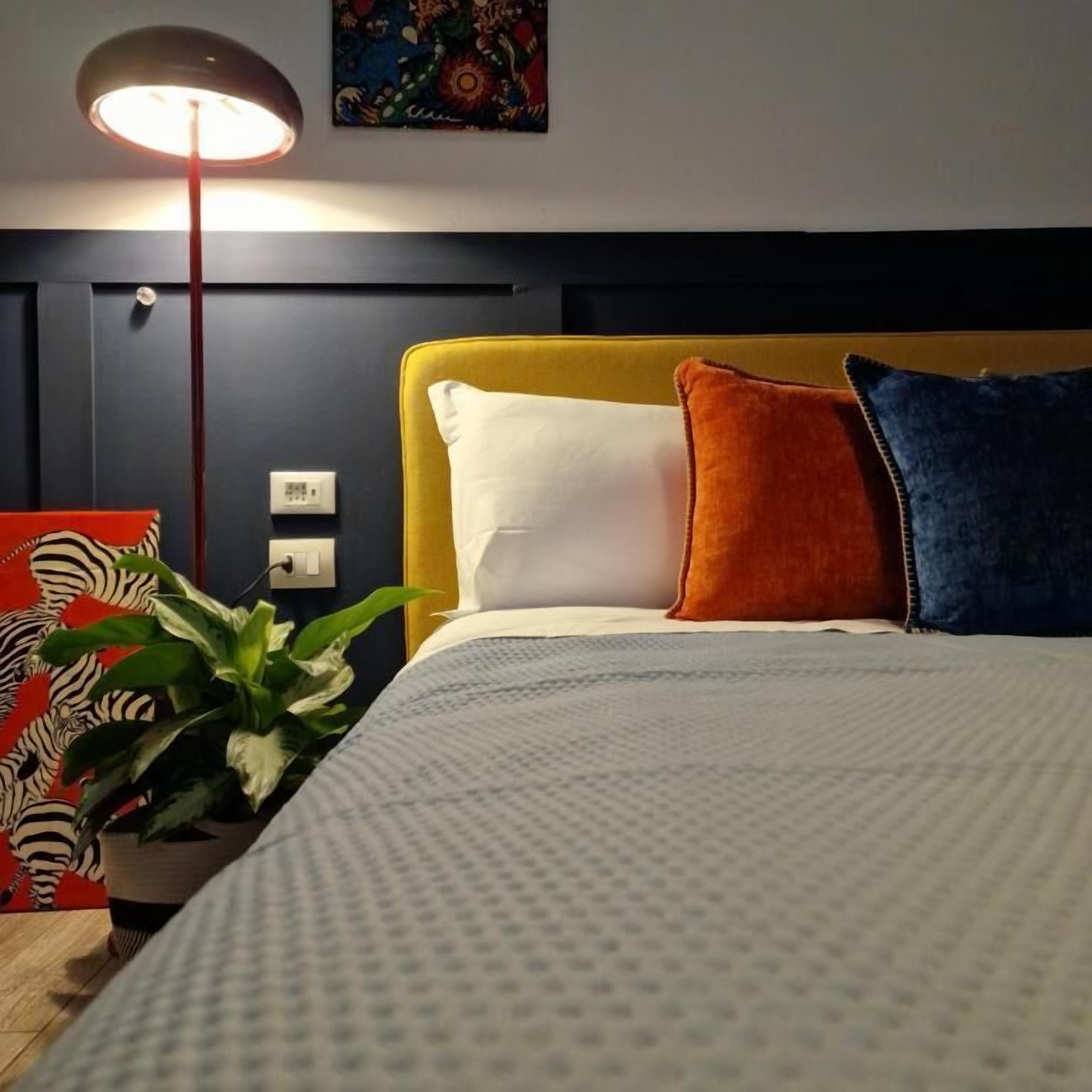 A neatly made bed with a yellow headboard, white and orange pillows, and a blue pillow. There is a potted plant on the side and a wall-mounted light fixture above the bed.