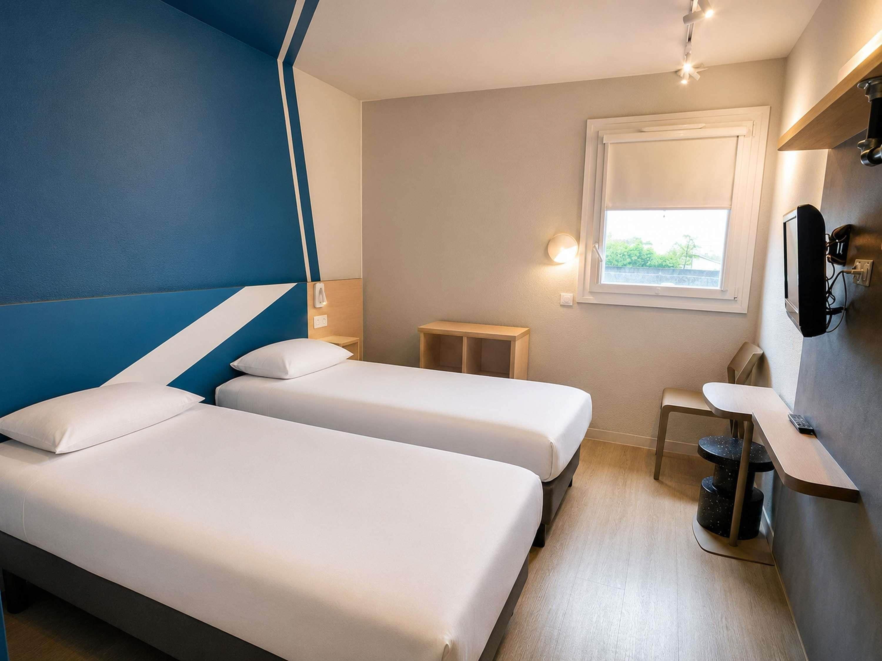 A hotel room with two beds, a small table, a chair, a TV, and a window.